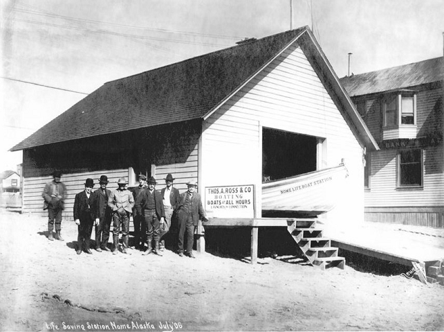 A July 1906 photograph showing the original boathouse and single bay entrance door for surfboat storage. The person standing second from the right is Keeper Thomas Ross, with other men that served in the initial station crew. (University of Washington digital archive)
